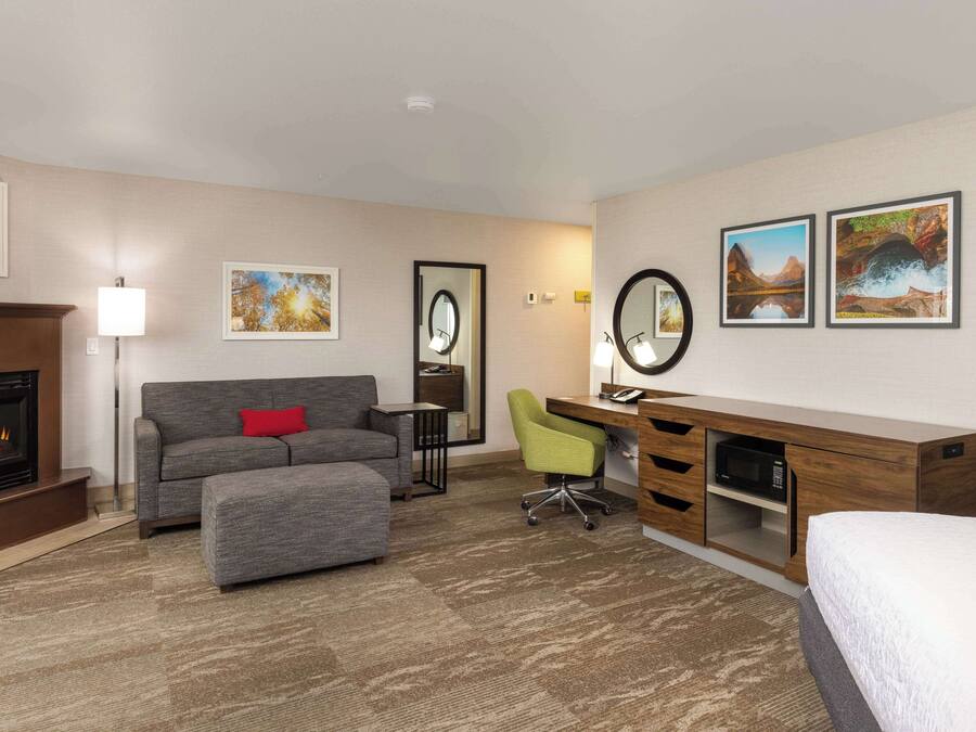 A hotel room with a fireplace, grey sofa, ottoman, desk with chair, and artwork on the walls.