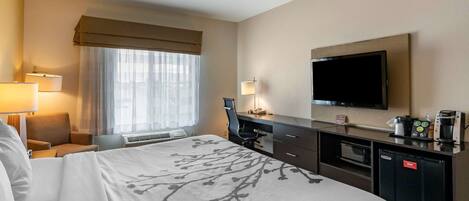 A hotel room with a large bed, a desk with a flat-screen TV, a coffee maker, and a mini-fridge.