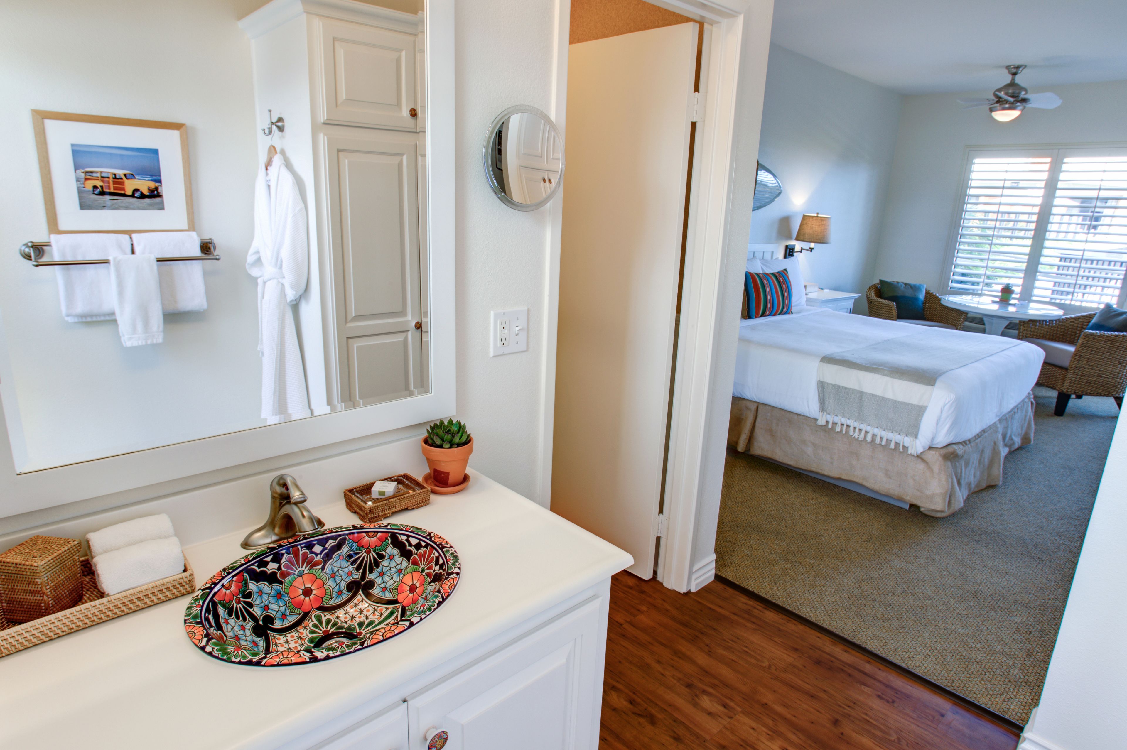 Habitación de hotel con baño que incluye un lavabo, espejo y barra para toallas. La habitación tiene una cama, una mesita pequeña y una ventana con persianas.