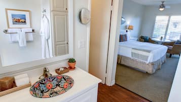 Habitación de hotel con baño que incluye un lavabo, espejo y barra para toallas. La habitación tiene una cama, una mesita pequeña y una ventana con persianas.