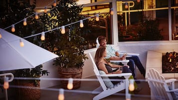 Un couple est assis sur des chaises blanches, sur une terrasse extérieure décorée de guirlandes lumineuses, d’un foyer et de plantes en pot.