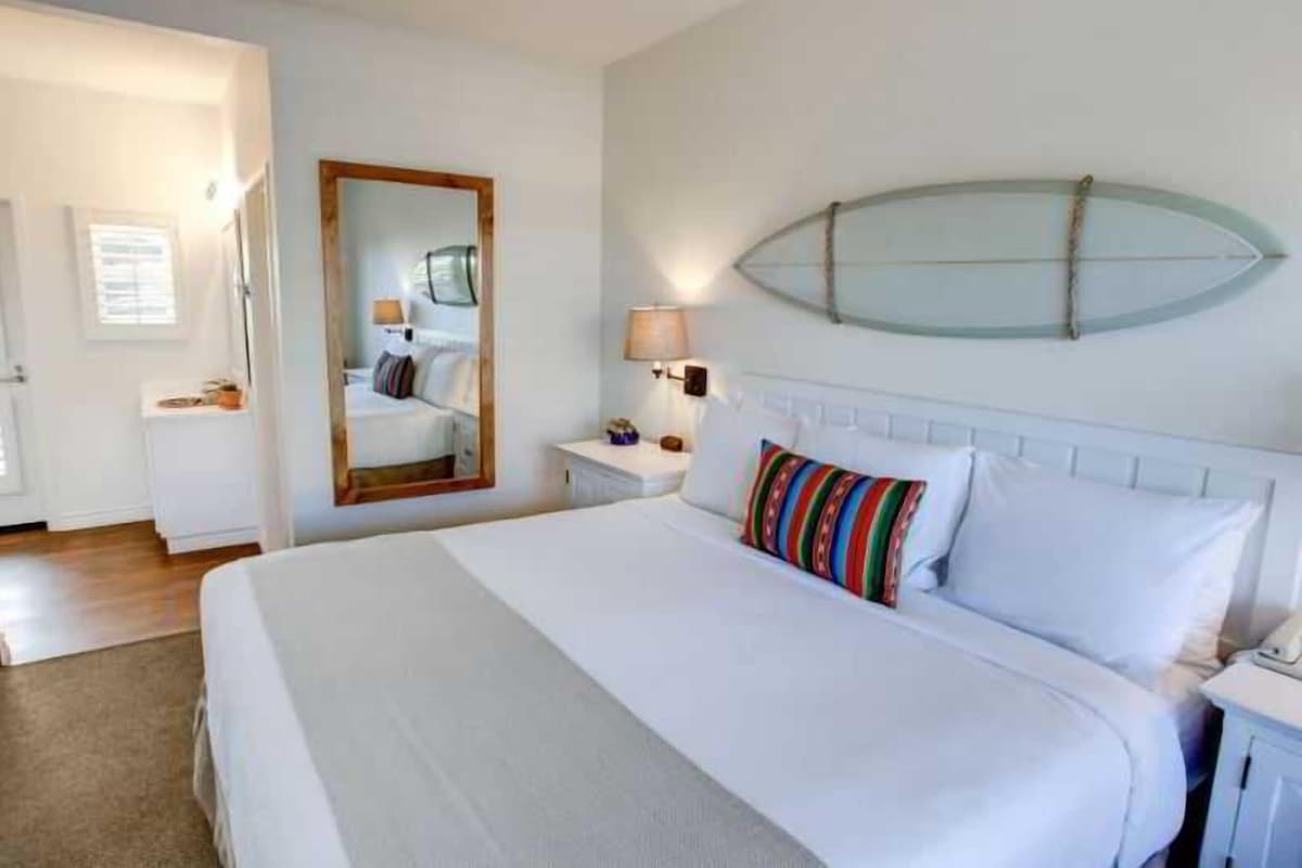 A hotel room with a large bed, a bedside table with a lamp, a mirror, and a surfboard decoration on the wall.
