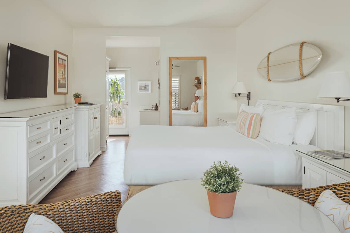 Quarto com cama, mesas de cabeceira, televisão, planta em vaso e vista para o exterior.