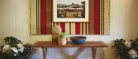 A wooden table with a framed picture, a woven ball, and a bowl. There are two potted plants on either side of the table.