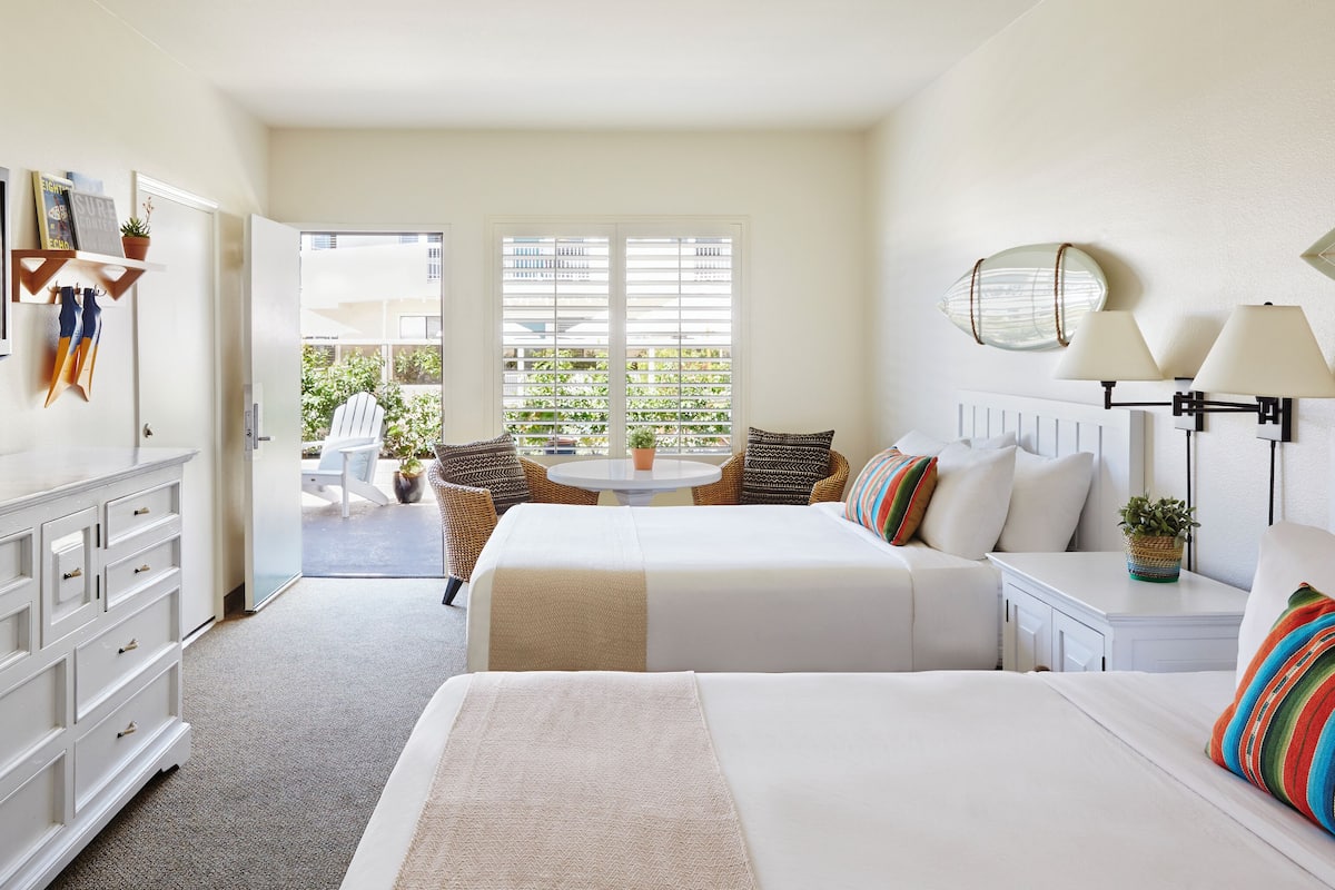 Quarto de hotel com duas camas, televisão, secretária com candeeiro e varanda com mobiliário exterior.