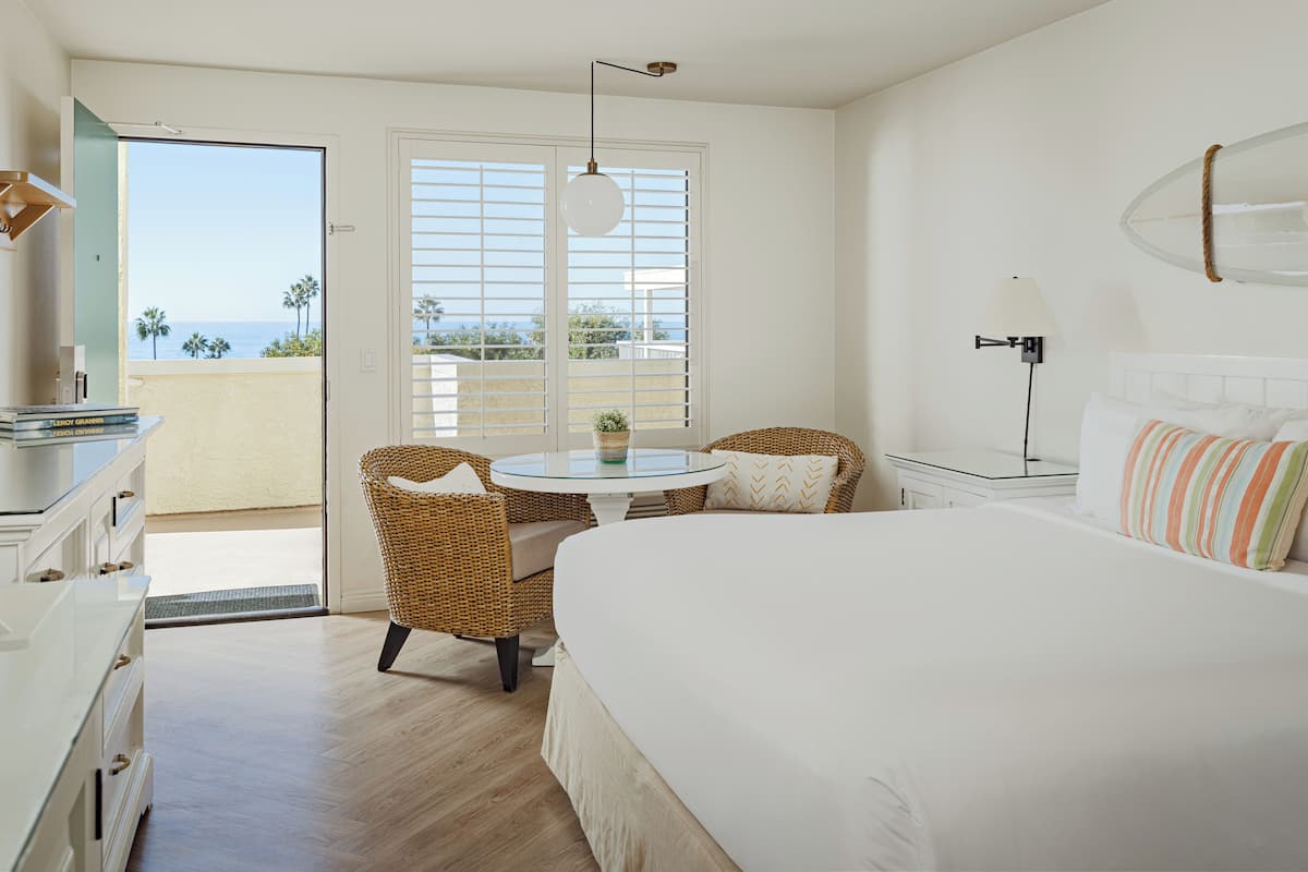 Quarto com cama, cômoda, mesa redonda com cadeiras, varanda com vista para o oceano e luminária pendente.