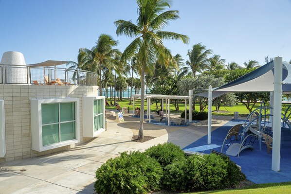 A playground with slides and swings, surrounded by palm trees and a covered seating area.