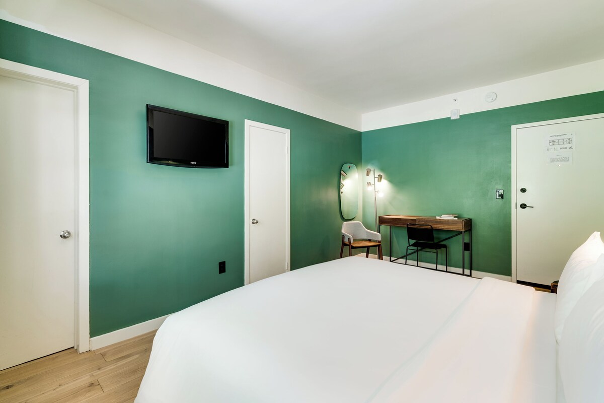A bedroom with a bed, a desk with a chair, a TV, and a door.