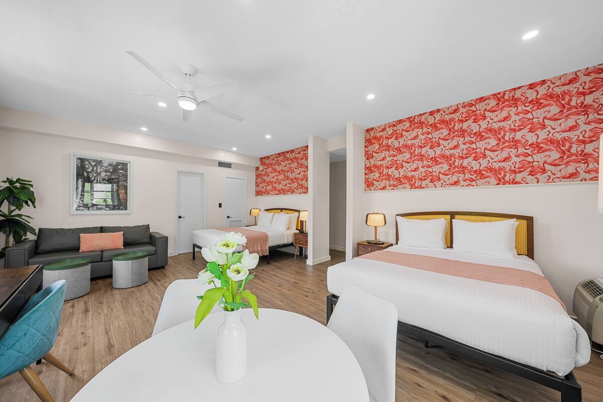 A hotel room with two beds, a sofa, a dining table with a vase of flowers, and a ceiling fan.