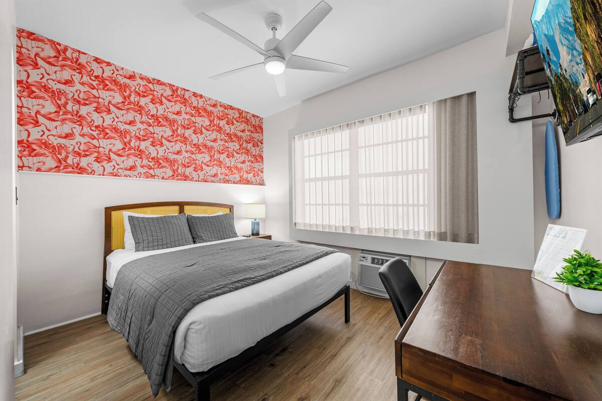 A bedroom with a bed, a ceiling fan, a window with blinds, a desk with a plant, and a wall with a red flamingo wallpaper.