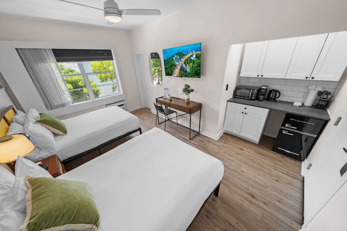A modern hotel room with two beds, a desk, a TV, and a kitchenette.