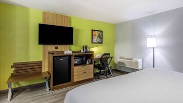 A hotel room with a bed, a desk with a computer, a small fridge, a microwave, a bench, a TV, and a painting on the wall.