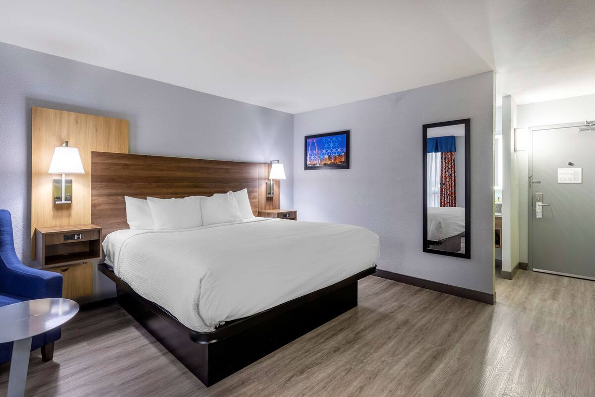 Een hotelkamer met een groot bed, nachtkastjes met lampen, een blauwe stoel en een spiegel aan de muur.