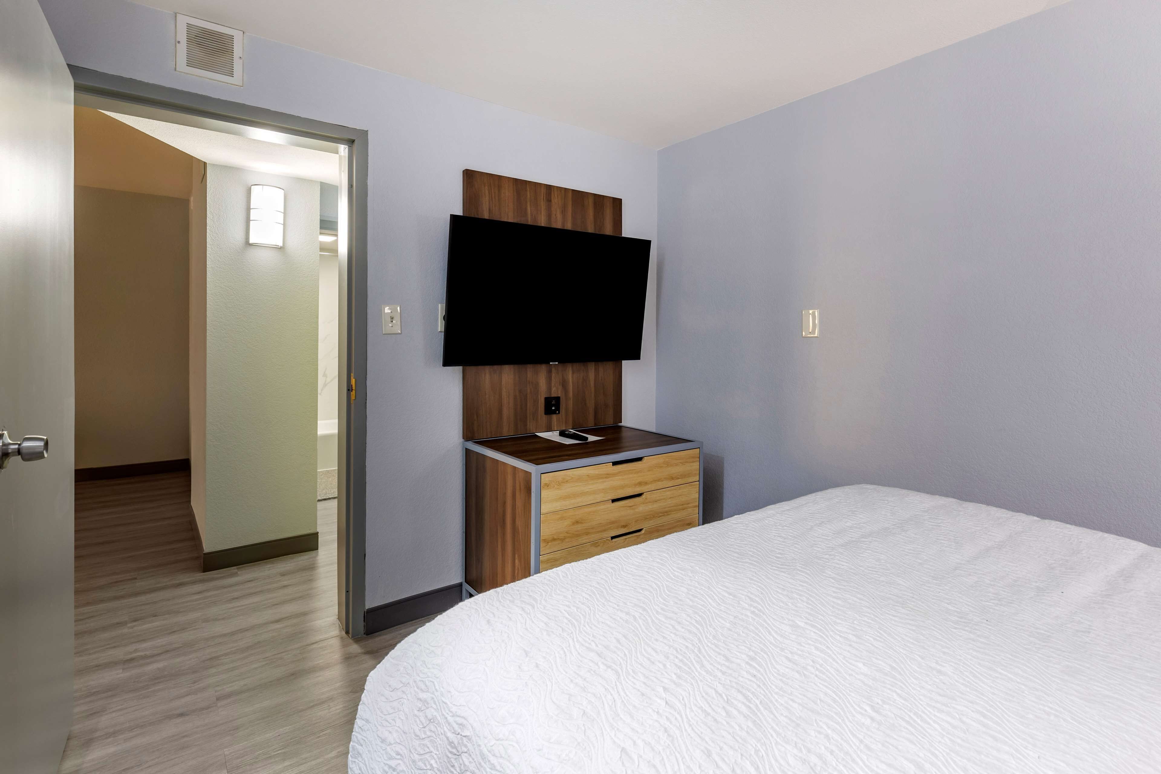 A hotel room with a bed, a wooden nightstand with a flat-screen TV, and a door leading to another room.