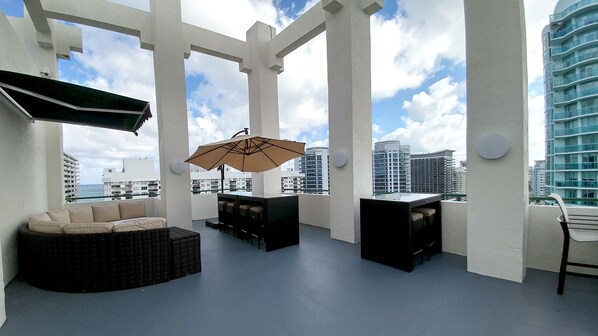 A rooftop terrace with a sofa, chairs, and an umbrella, overlooking a cityscape with tall buildings.