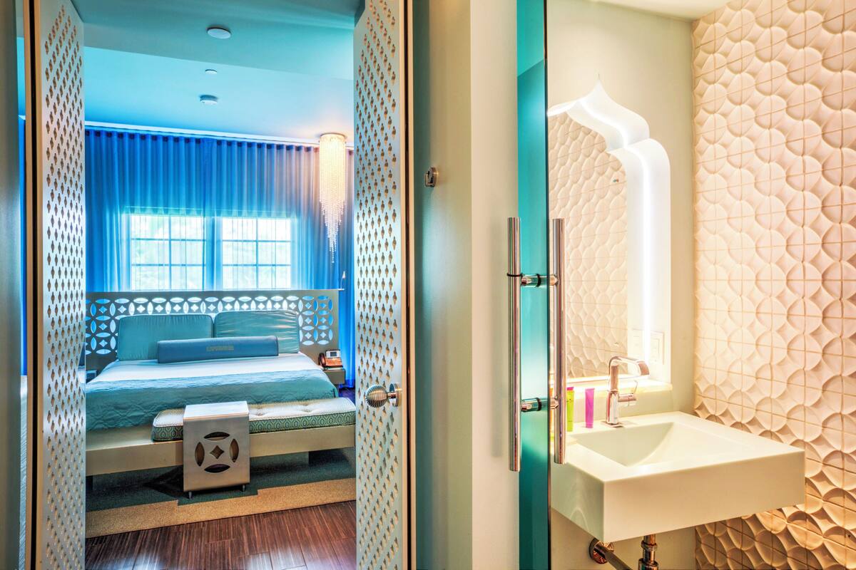 A hotel room with a bed, a bedside table, a window with curtains, and a bathroom with a sink and mirror.