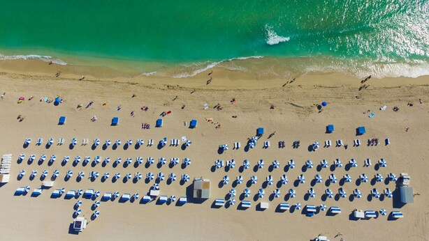 An aerial view of a beach with rows of beach umbrellas and sun loungers, a sandy shore, and waves approaching the beach.