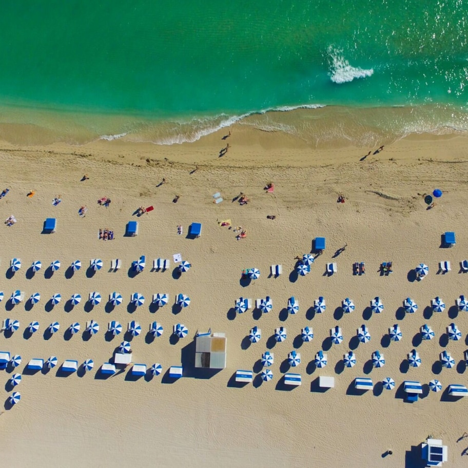 An aerial view of a beach with rows of beach umbrellas and sun loungers, a sandy shore, and waves approaching the beach.