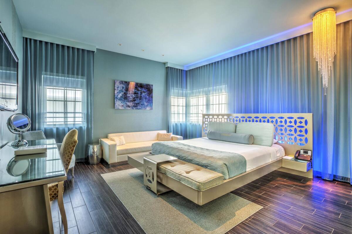 A modern hotel room with a large bed, a desk with a chair, a small table, and a decorative wall panel.