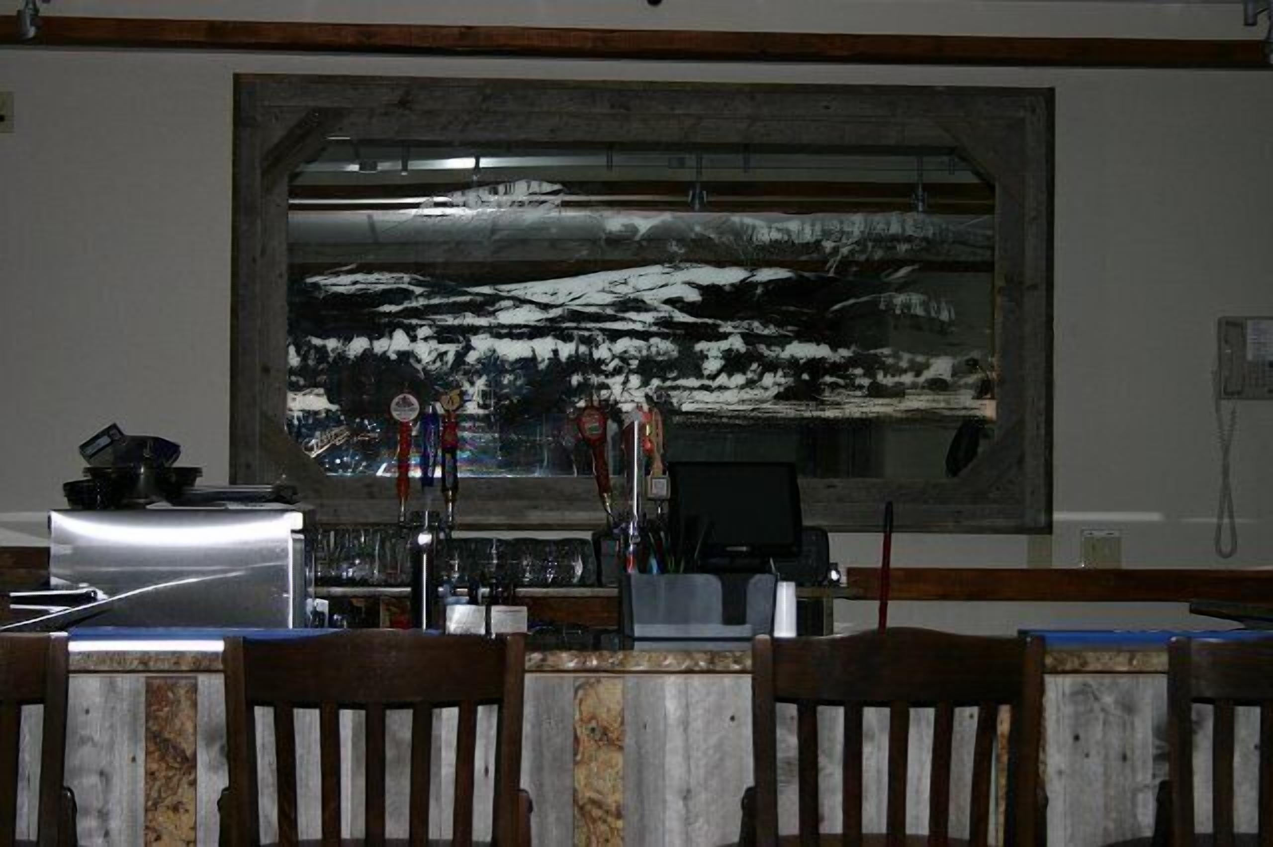 A bar area with a large painting of a snowy landscape on the wall, wooden chairs, and a counter with various items.