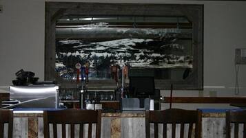 A bar area with a large painting of a snowy landscape on the wall, wooden chairs, and a counter with various items.