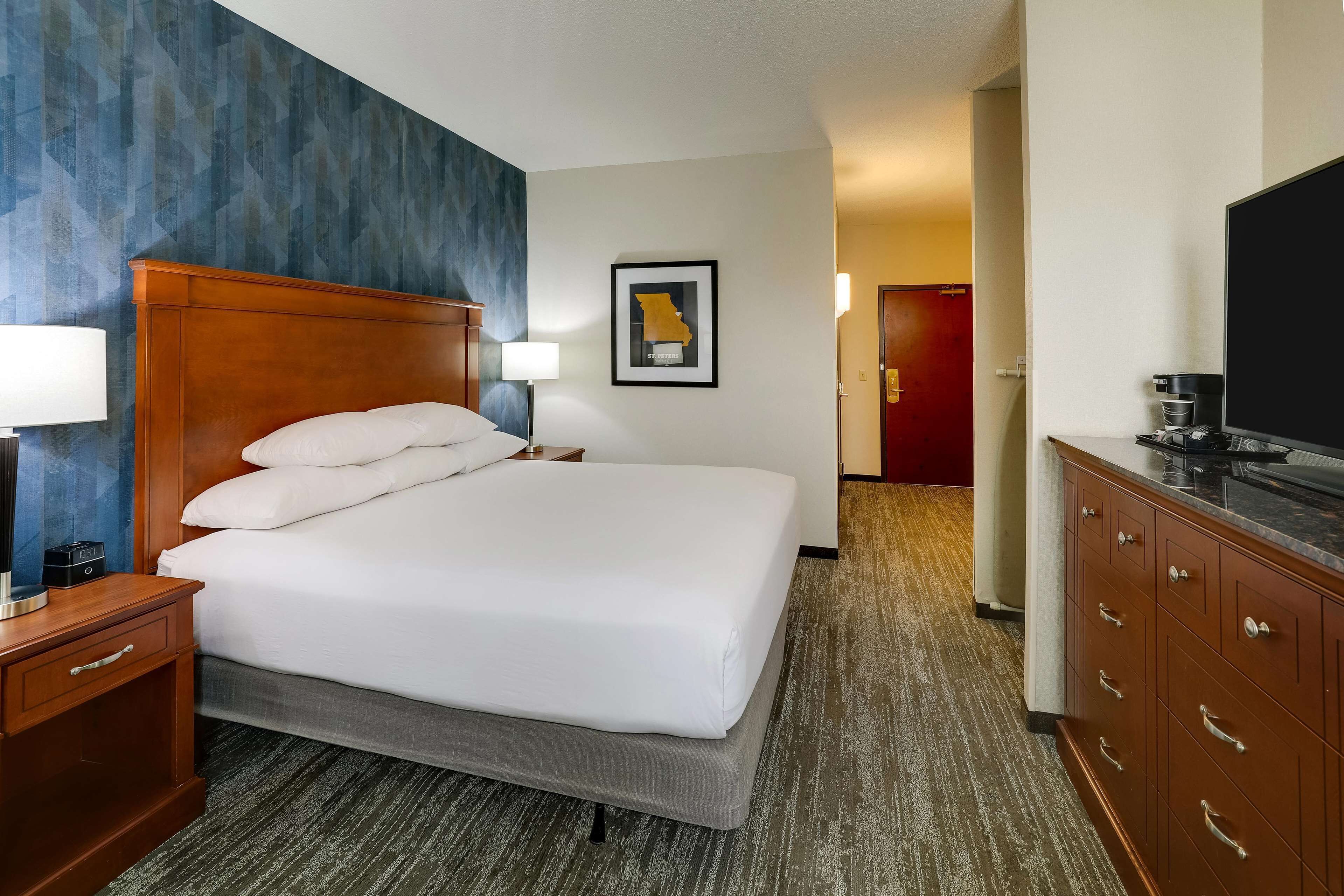 A hotel room with a large bed, a wooden nightstand with a lamp, a framed map on the wall, and a television on a cabinet.