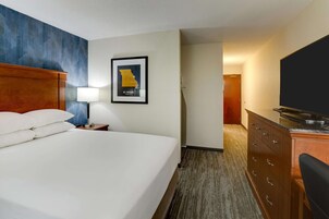 Quarto de hotel com uma cama grande, cabeceira de madeira, escrivaninha com abajur, televisão de tela plana, mapa emoldurado na parede e piso coberto por carpete.