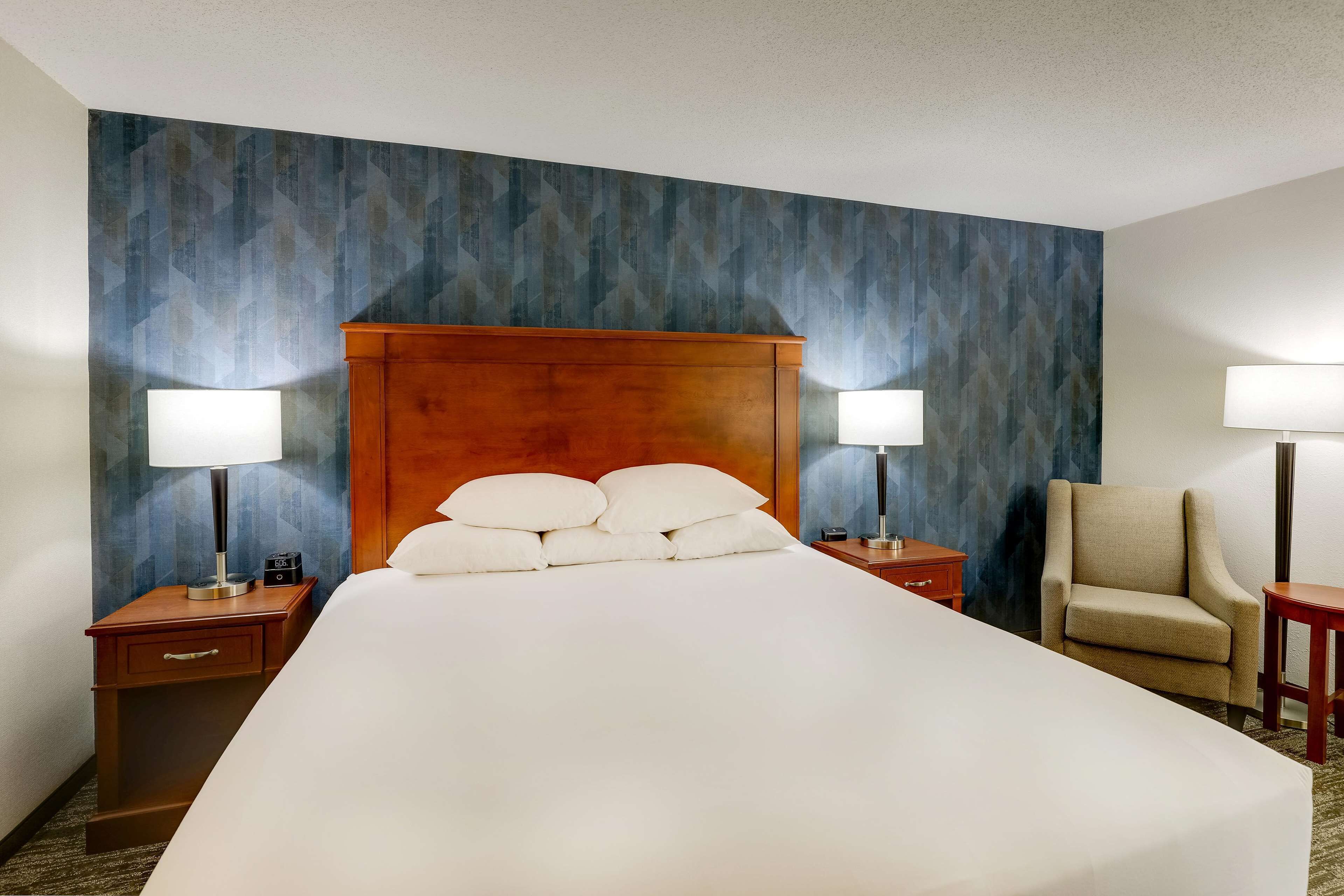 Habitación de hotel con una cama grande, dos mesitas de noche con lámparas, un cabecero de madera, una silla y una pared con un diseño geométrico.