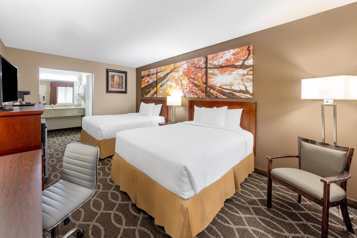 A hotel room with two beds, a desk, a chair, a TV, and a large painting on the wall.