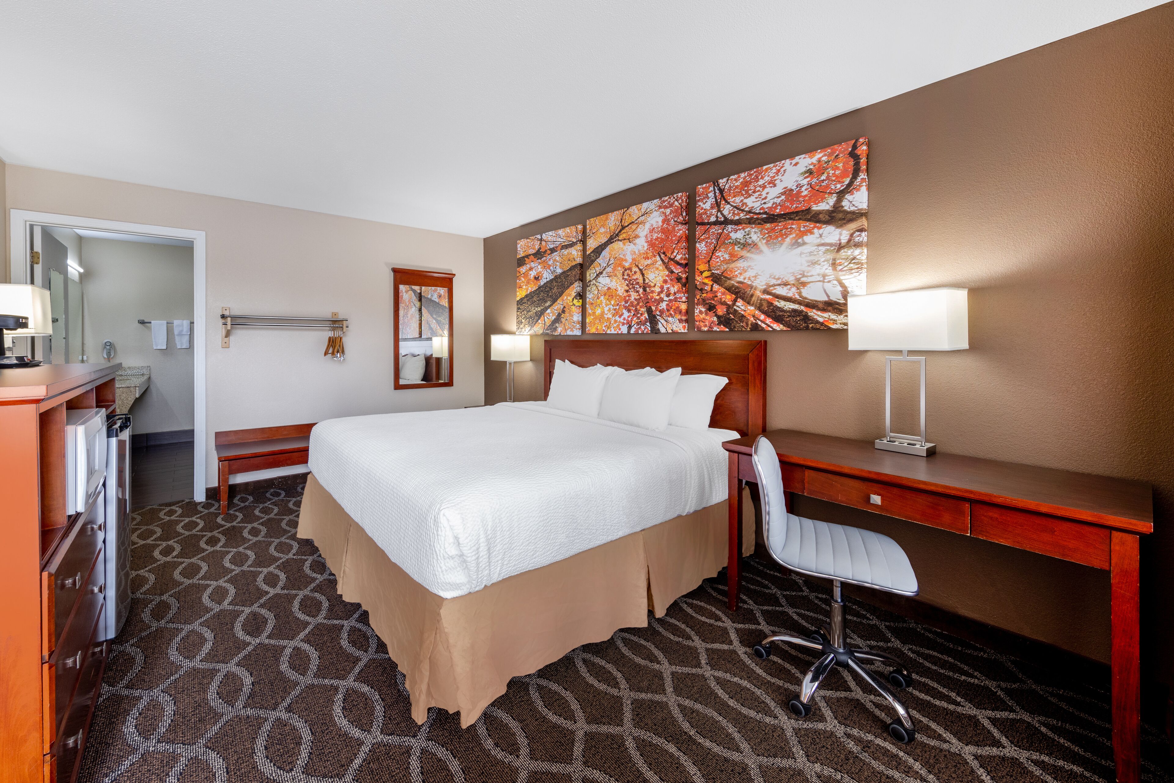 A hotel room with a large bed, a desk, a chair, a desk lamp, a mirror, and a painting on the wall.
