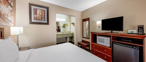 Habitación de hotel con cama, televisión, microondas, refrigerador, cómoda de madera, espejo y baño con lavamanos y espejo.