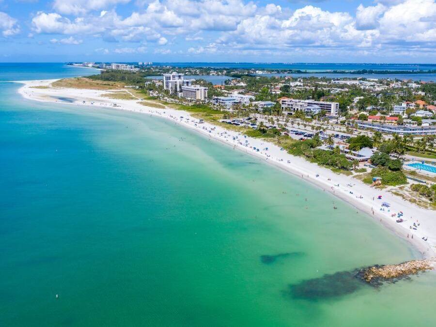 An aerial view of a coastal resort with a long sandy beach, clear turquoise waters, and a mix of residential and hotel buildings.