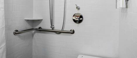 Un baño con paredes de azulejos blancos, una ducha con cabezal de mano, un asidero y un lavamanos blanco con espejo.