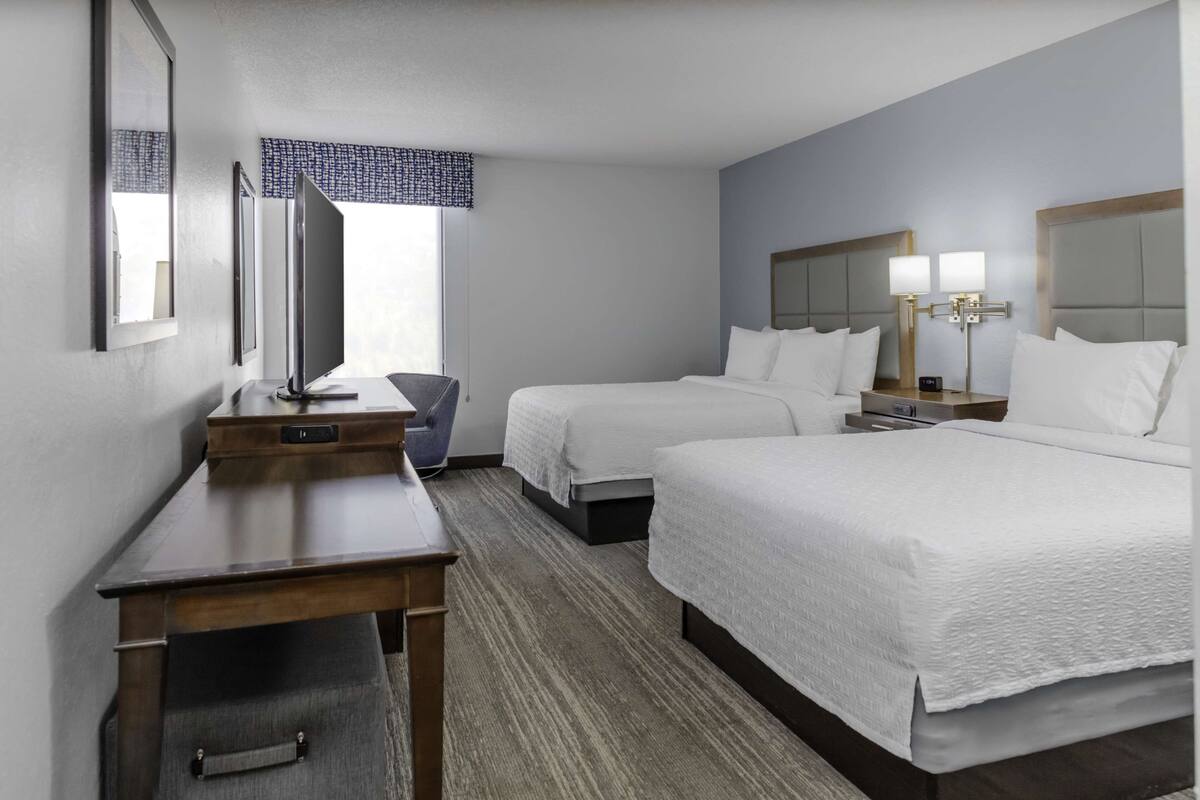 A hotel room with two beds, a desk, a chair, a TV, and a window with a patterned curtain.