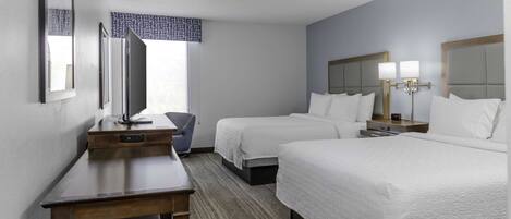 A hotel room with two beds, a desk, a chair, a TV, and a window with a patterned curtain.