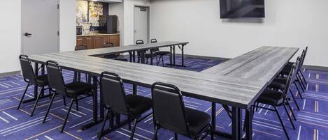 Un salón de conferencias con una larga mesa, sillas negras, un televisor y una zona de cocina al fondo.