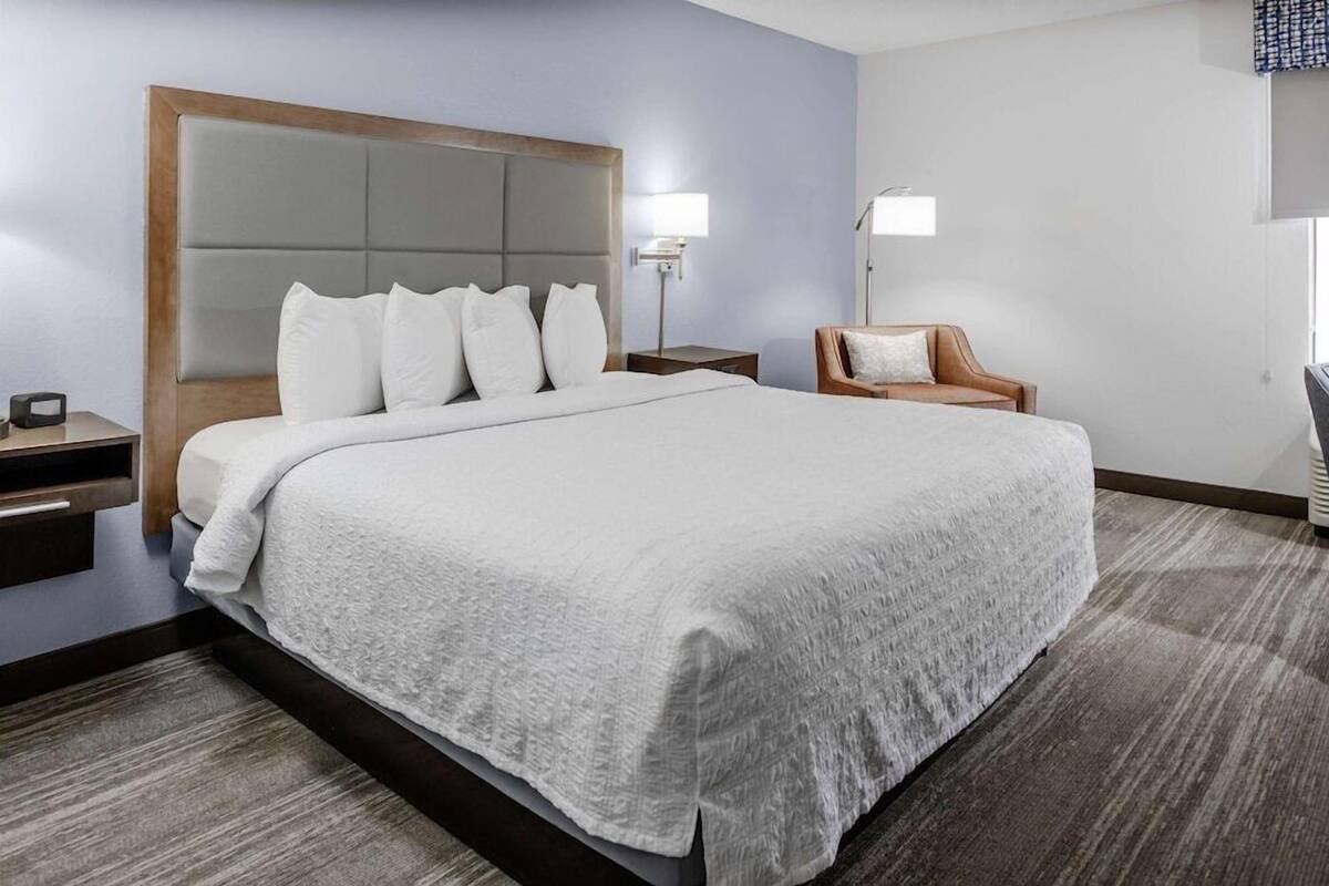 A hotel room with a large bed, a nightstand, a chair, a lamp, and a wall-mounted TV.