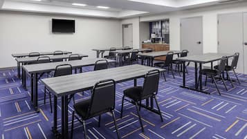 A conference room with multiple tables and chairs, a flat-screen TV mounted on the wall, and a kitchenette area.