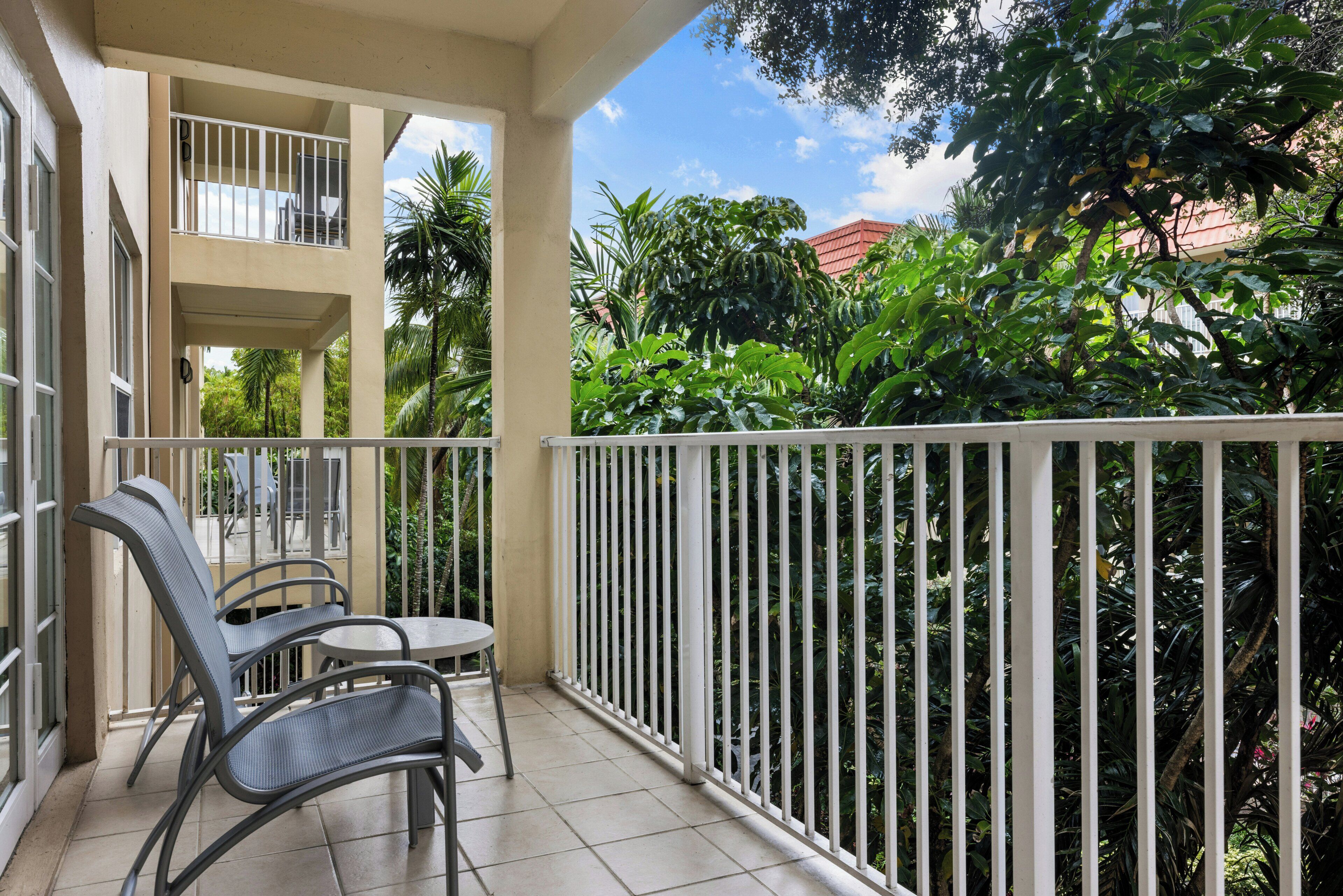 A balcony with two chairs and a table, surrounded by a white railing, overlooking lush greenery and a building with a red roof.