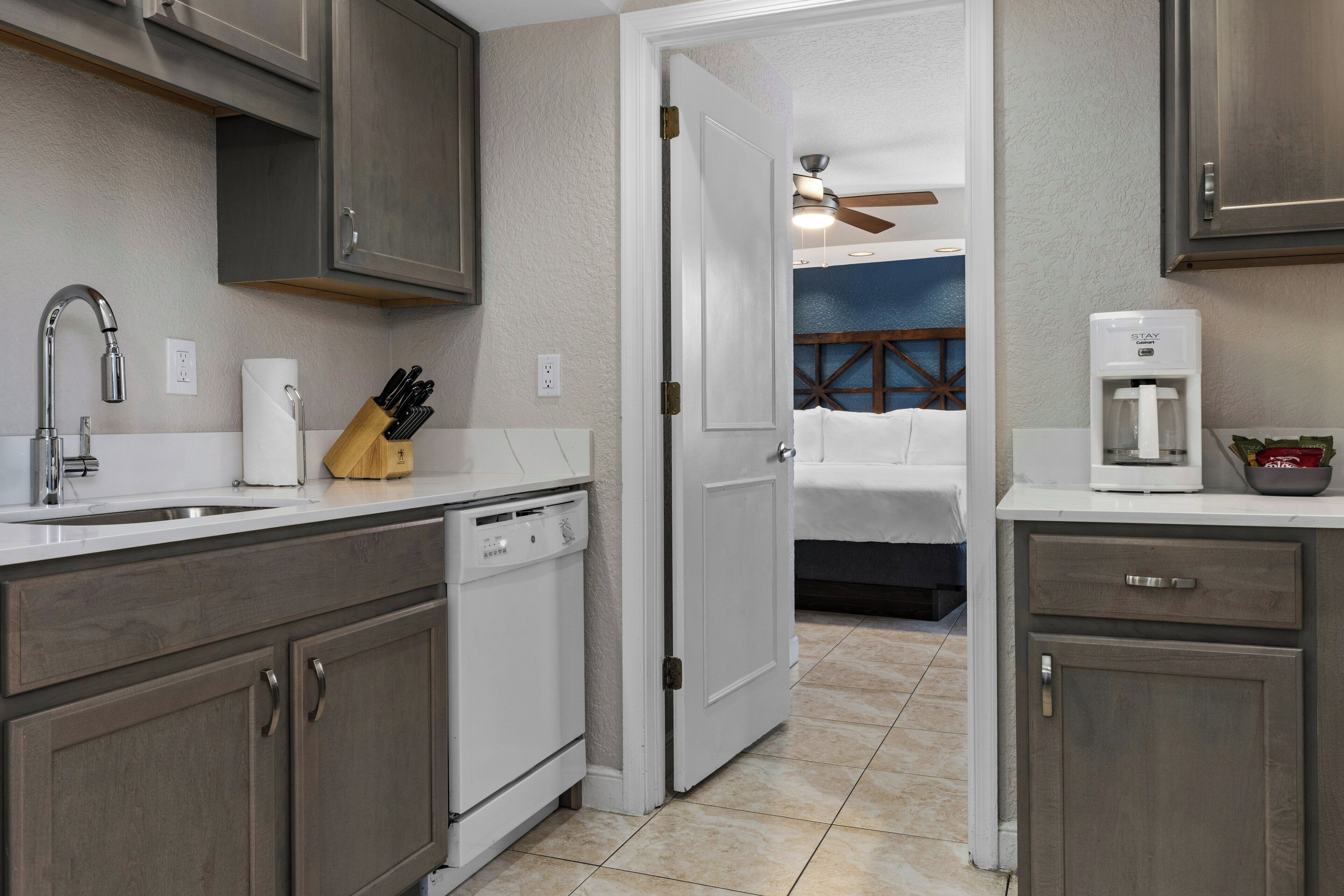 A kitchen with a dishwasher, a coffee maker, and a door leading to a bedroom.