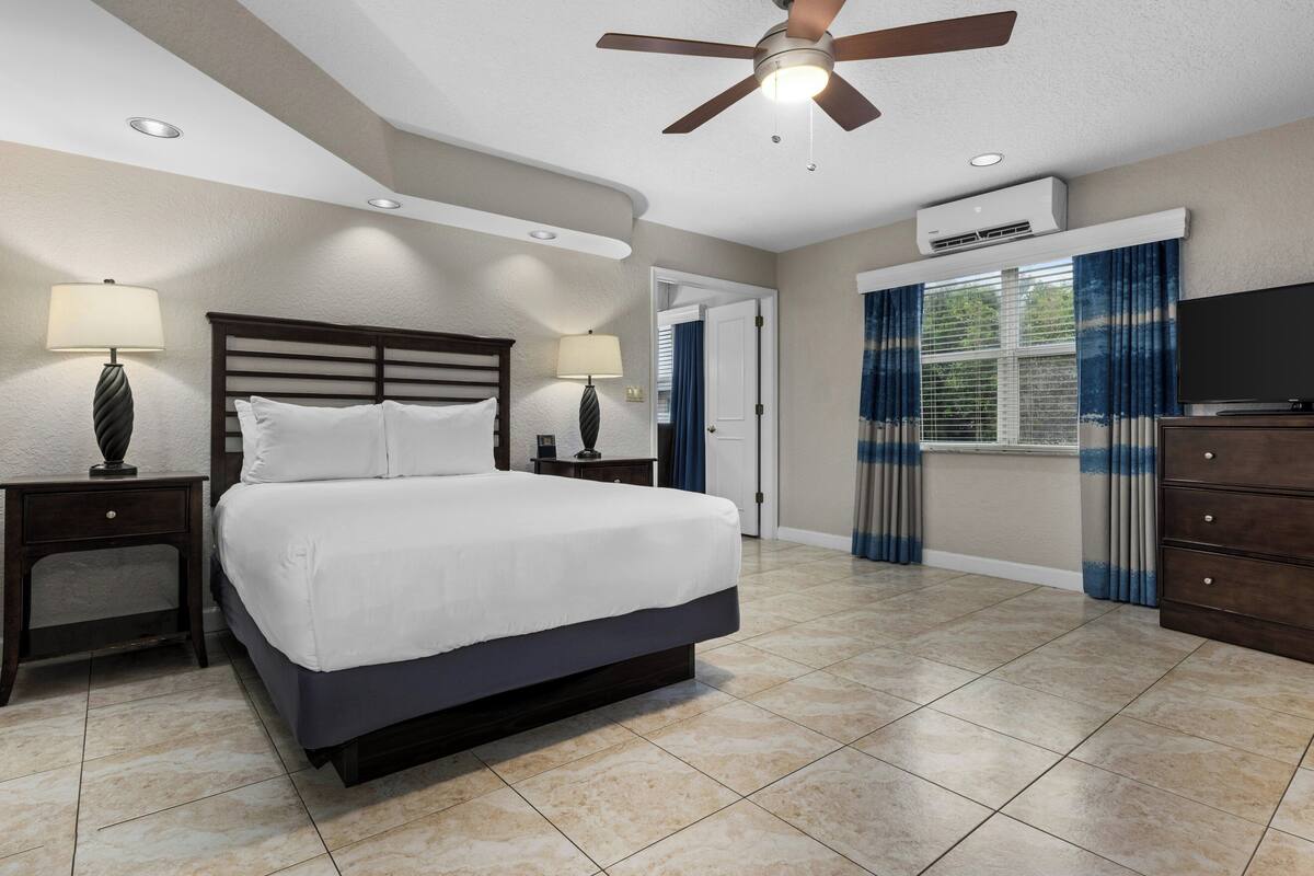 A hotel room with a bed, two bedside tables with lamps, a ceiling fan, an air conditioning unit, a dresser, and a television.