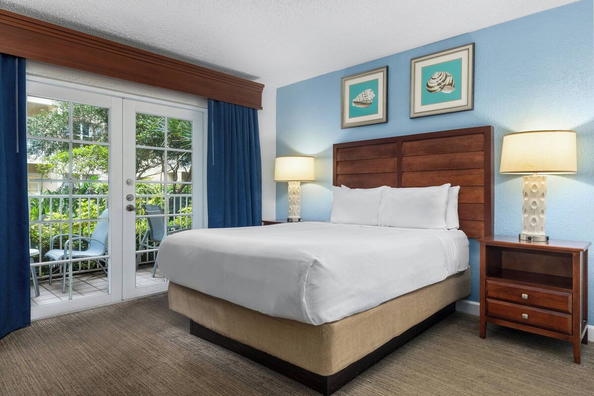 A hotel room with a large bed, two bedside lamps, a wooden nightstand, and a view through a glass door to an outdoor seating area.