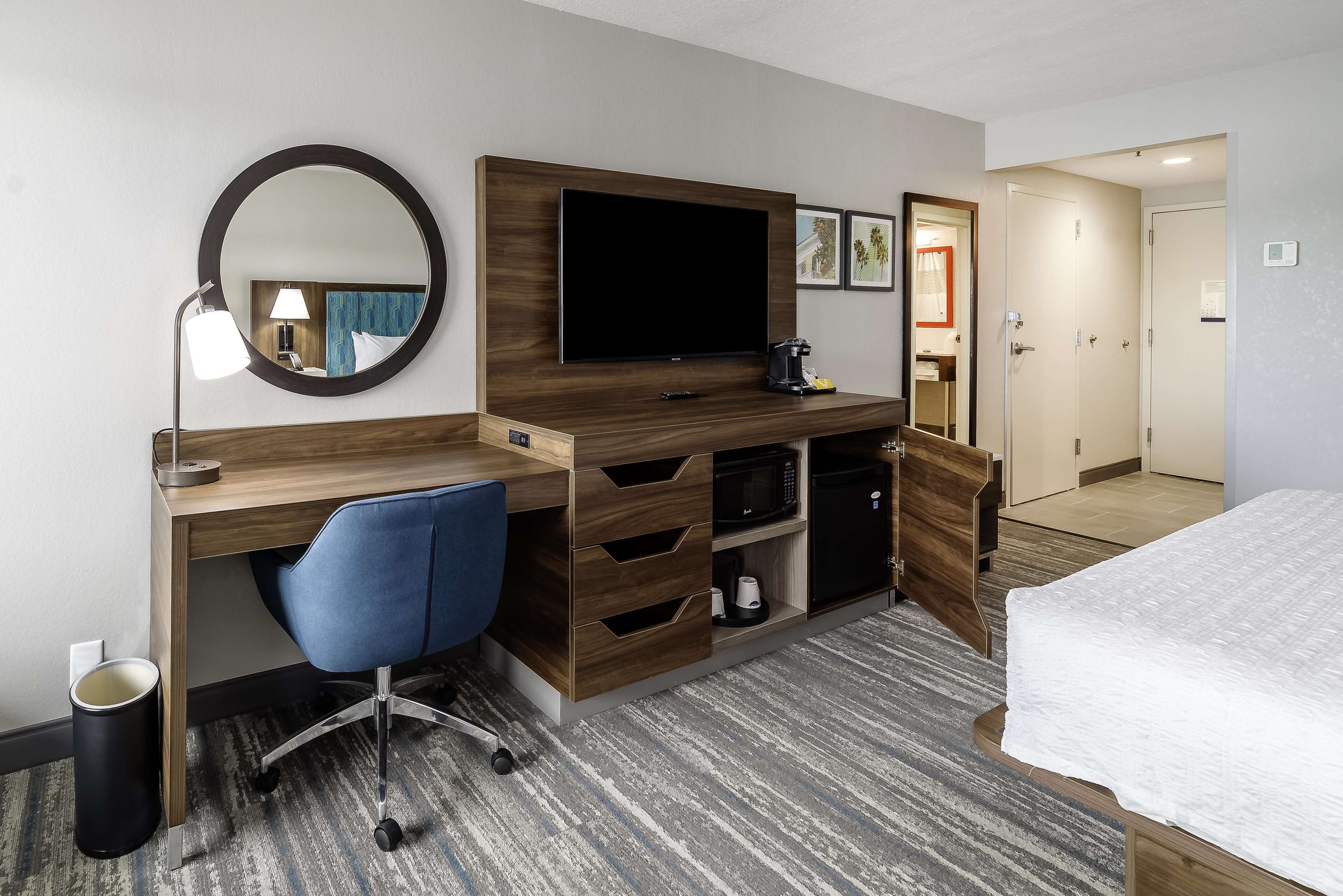 A hotel room with a wooden desk, a blue office chair, a flat-screen TV, a coffee maker, a microwave, and a bed with a white comforter.