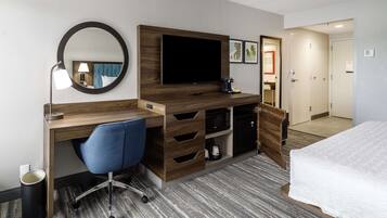 A hotel room with a wooden desk, a blue office chair, a flat-screen TV, a coffee maker, a microwave, and a bed with a white comforter.