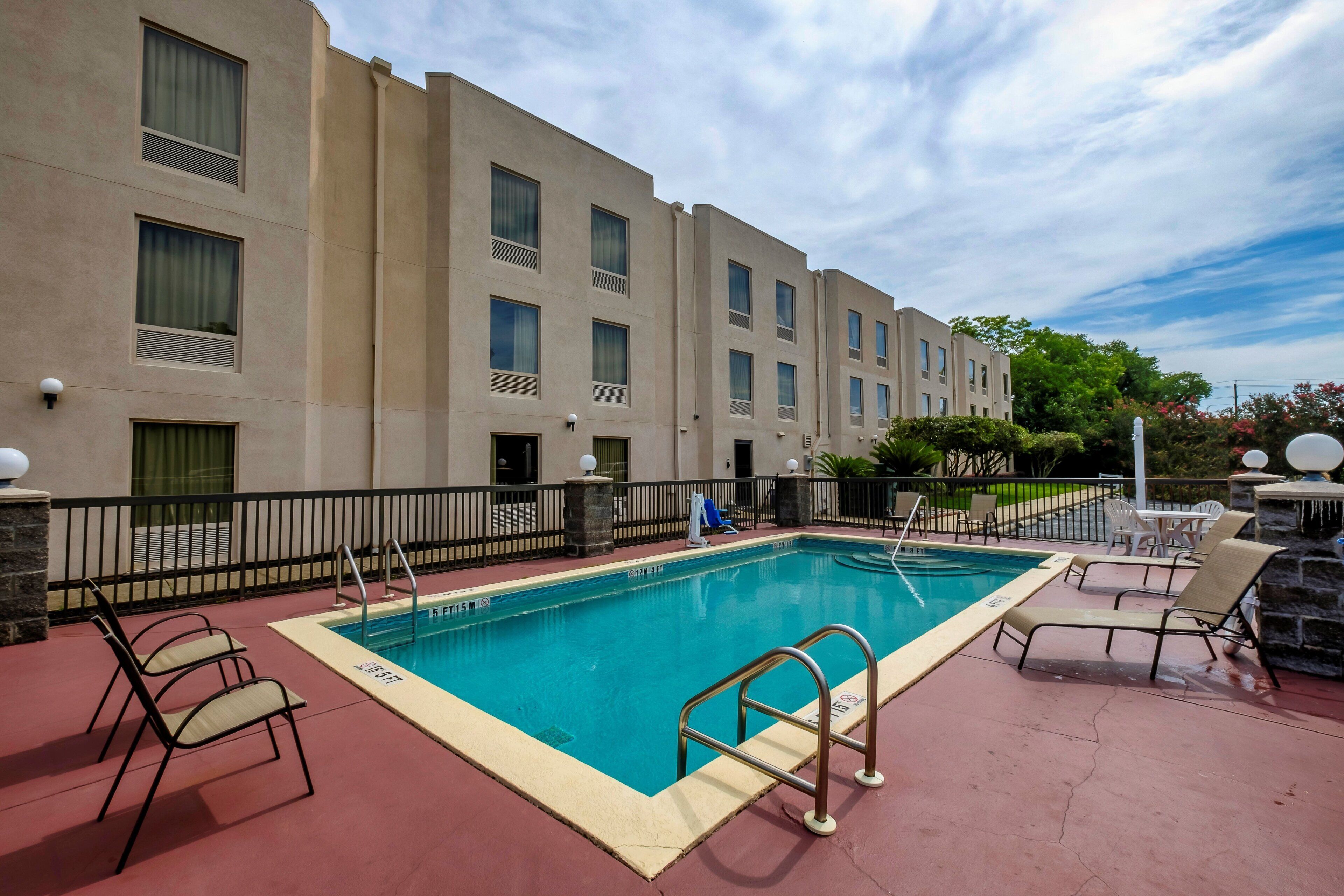 A hotel pool area with a rectangular pool, lounge chairs, and a building with multiple windows and balconies.