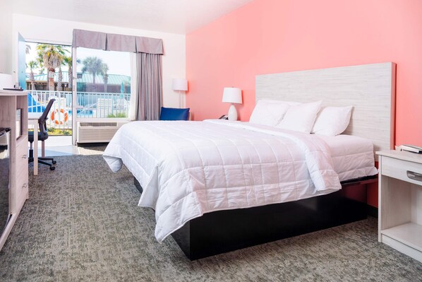 A hotel room with a bed, a desk, a chair, a nightstand, and a view of a pool area.
