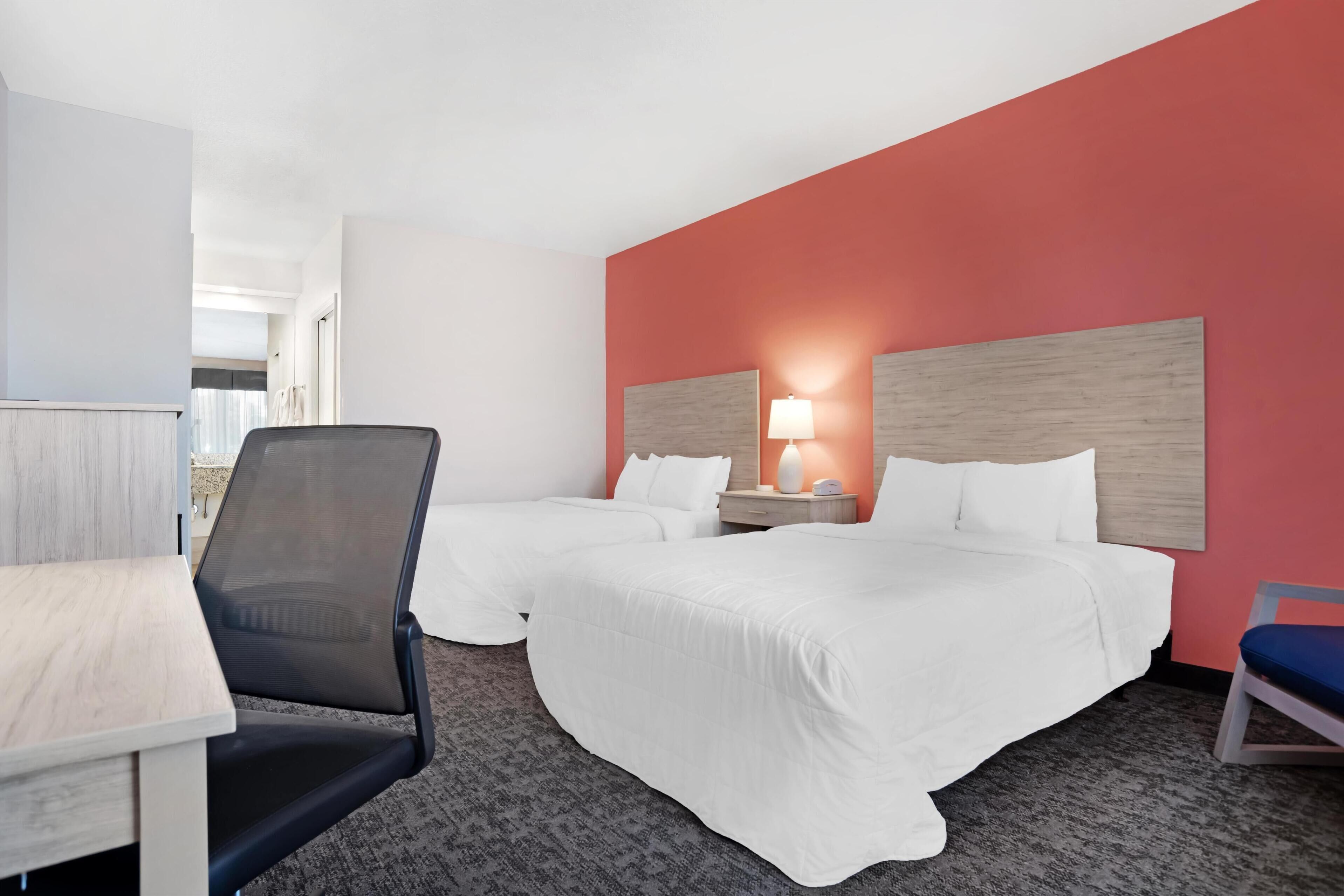 A hotel room with two beds, a desk, a chair, and a nightstand with a lamp.