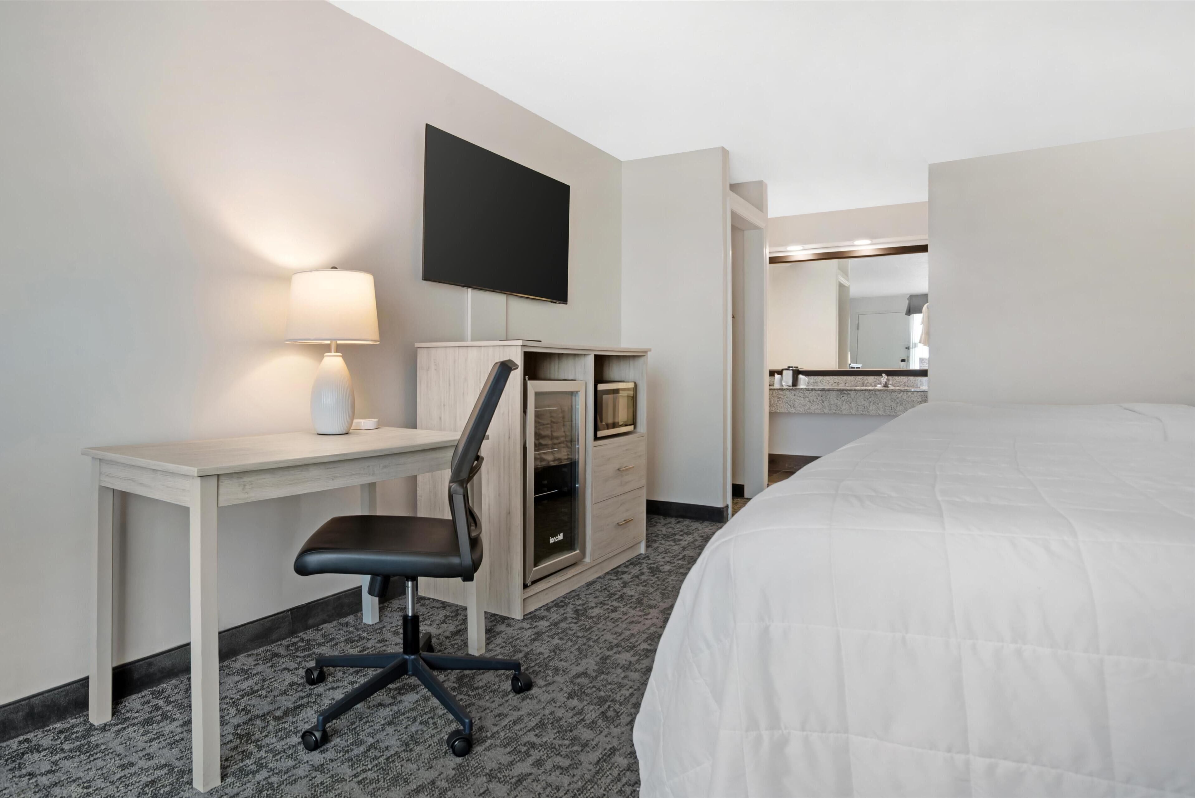 A hotel room with a bed, a desk with a chair, a flat-screen TV, a lamp, and a minibar.