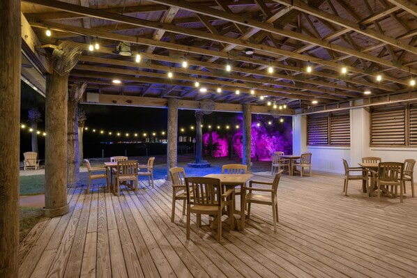 An outdoor wooden deck with tables and chairs, string lights, and a covered area with a wooden ceiling.