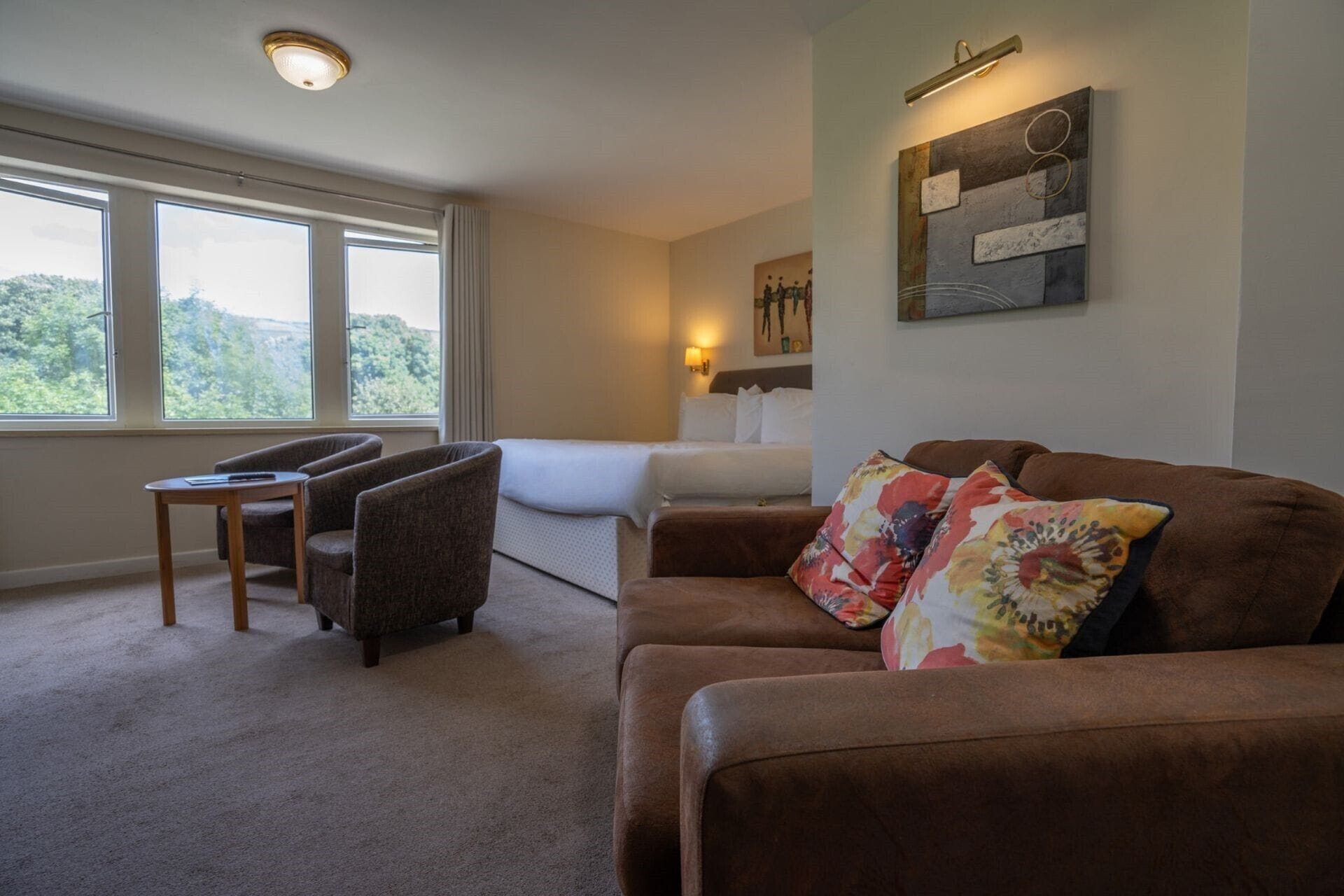 A hotel room with a bed, a brown sofa, a small table, and a window with a view of trees.